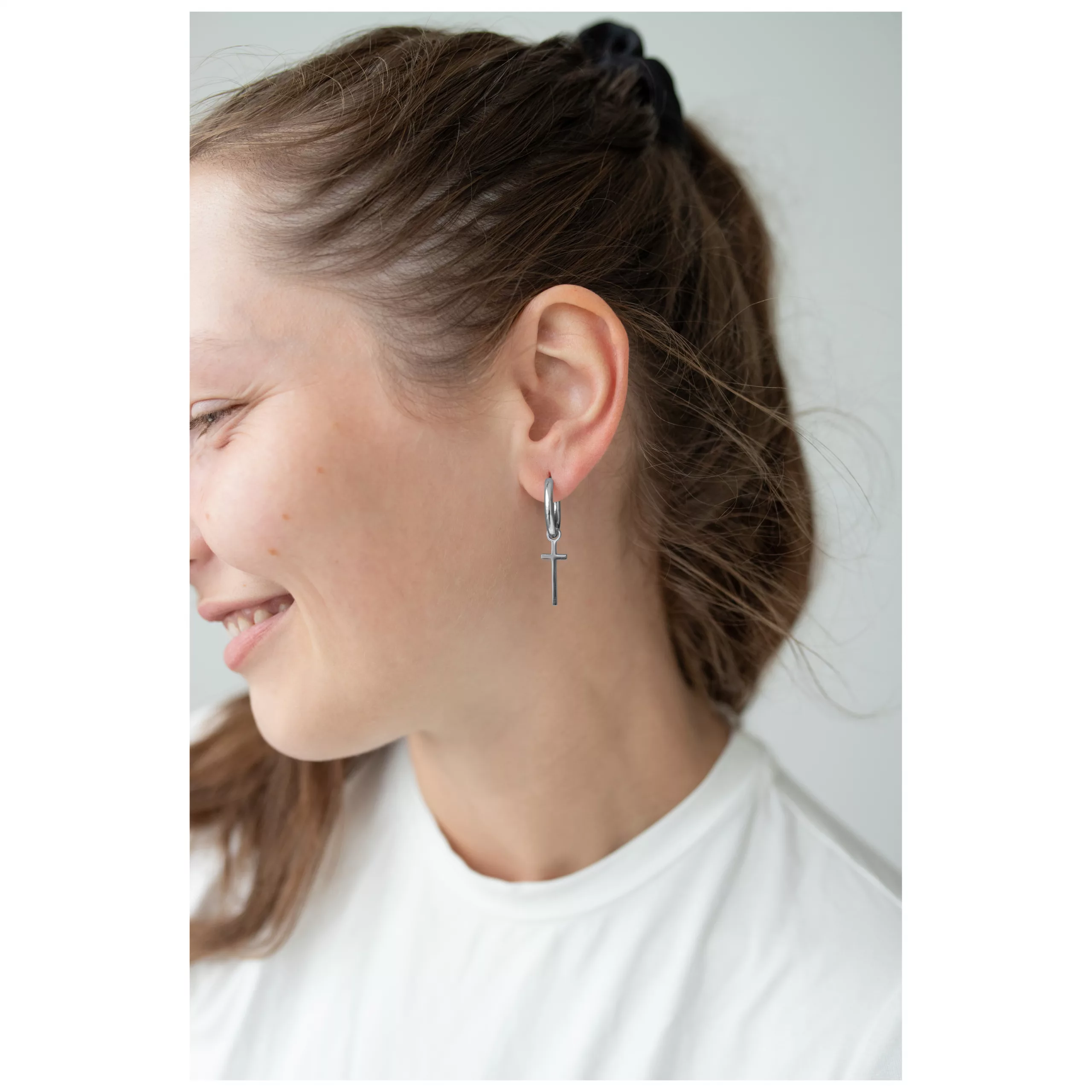 Elegante Little Cross Hoops in Silber Frau trägt silberne Kreuz-Ohrringe, lächelnd im Profil auf hellem Hintergrund