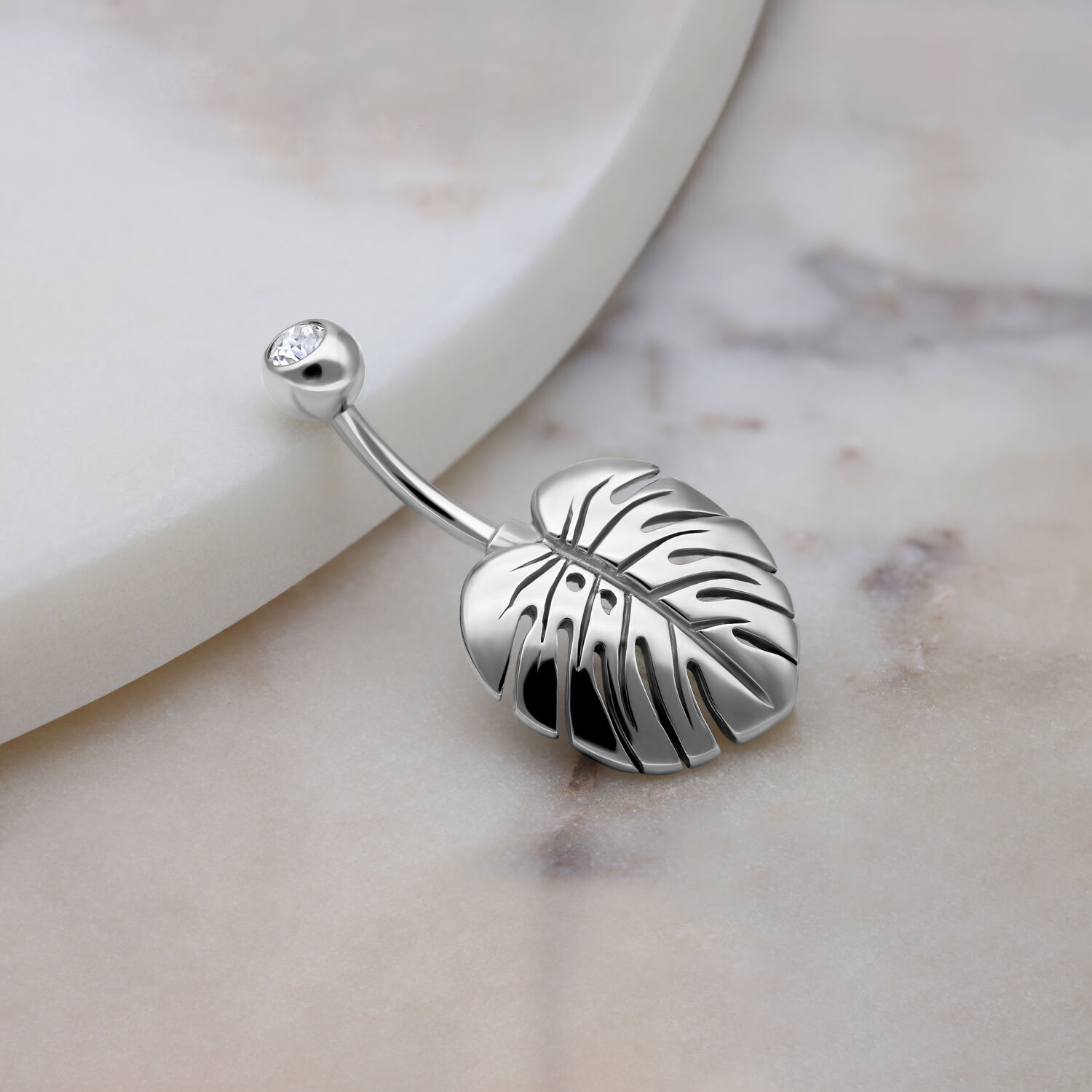 Monstera Blatt Bauchnabelpiercing Silber Silbernes Bauchnabelpiercing mit Monstera Blatt und Kristall auf Marmorhintergrund