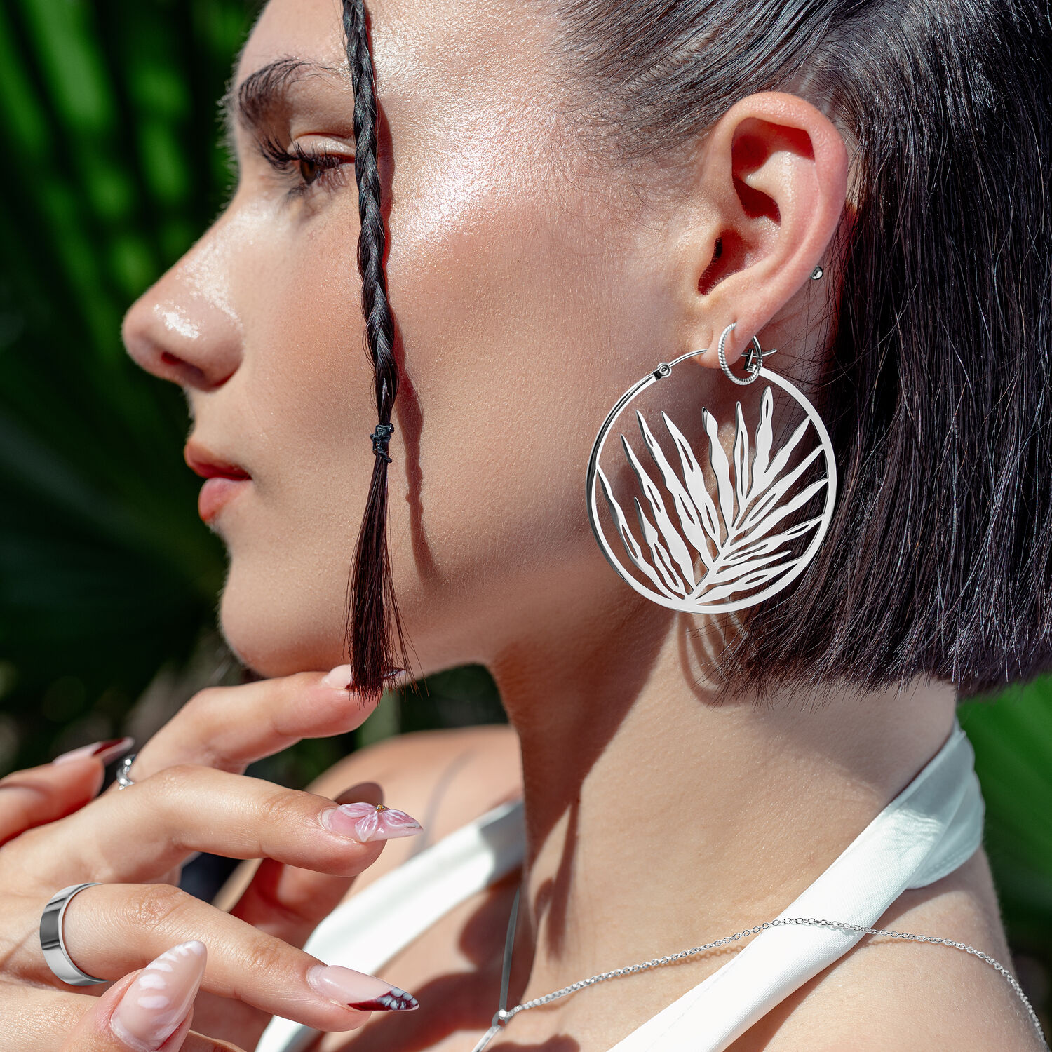 Noble Leaf Hoops in Silber an Ohr Silberne Noble Leaf Hoops Ohrringe an Frau mit geflochtenem Haar und tropischem Hintergrund